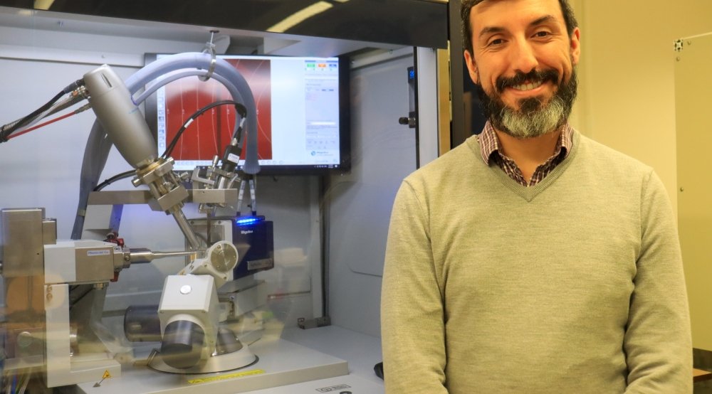 Smiling bearded man in a sweater stands before a scientific apparatus