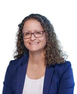 Upper body photo of a smiling person in glasses and with shoulder-length curly brown hair. They are wearing a blue business jacket, a white shell top, and a small pendant.