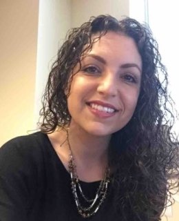 A smiling fair-skinned woman with long, dark, curly hair, a necklace, and a black top