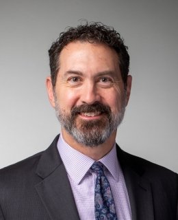 Smiling light-skinned man with salt-and-pepper beard, dark hair, lilac shirt, suit jacket, and tie