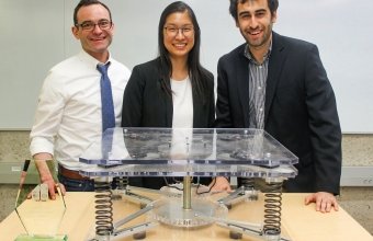 3 smiling people stand behind a pale veneer table on which rests a device made of clear plastic and metal.