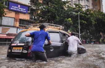 Two men push a small black car down a flooded street; the water reaches the tops of their legs.