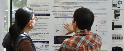 Two people stand before a scientific poster, their backs turned; one is pointing at it