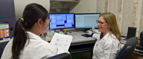 Two scientists examine a document while sitting in front of monitors