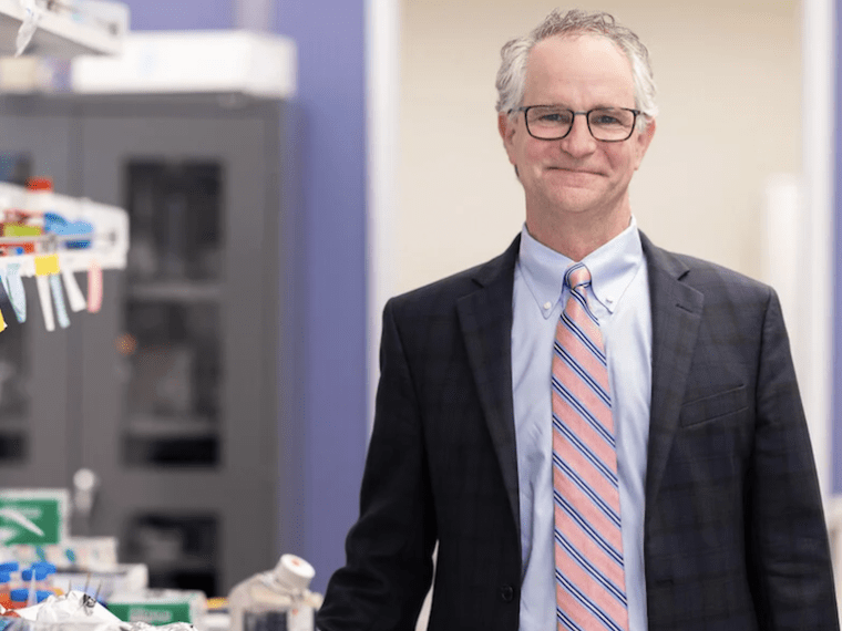 A smiling man in a suit and tie stands by a lab bench